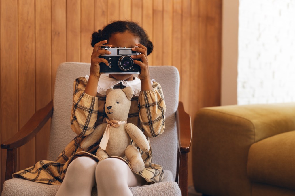 Vue d'une petite fille métisse avec des couettes habillée en robe. Elle est assise sur une chaise dans un décor rétro et tient un vieil appareil photo.