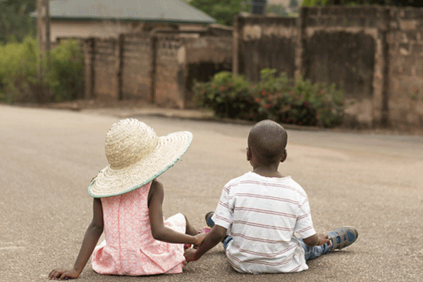 Vue de dos. Couple d'enfants noirs africains assis dans un village en Afrique et se tenant par la main. La petite fille porte une robe rose et un chapeau de paille.