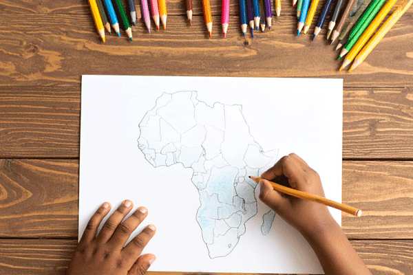 Vue du dessus d'un petit garçon qui dessine la carte de l'Afrique sur une feuille blanche posée sur une table en bois, et entourée de crayons de couleurs.