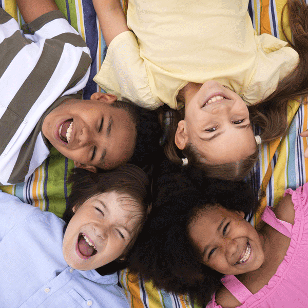 Vue du haut. Groupe d'enfants allongés sur le dos par terre dans un parc en train de rire. Leur tête se touche.